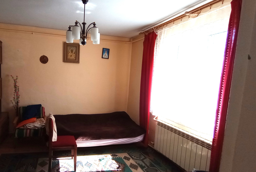Zdjęcie przedstawia tradycyjnie urządzone, jasne pomieszczenie mieszkalne o brzoskwiniowych ścianach. Po prawej stronie znajduje się duże okno, przez które wpada intensywne, naturalne światło dzienne. Okno przesłonięte jest wzorzystą, jasną firanką oraz ujęte w długie, kontrastujące, czerwone zasłony, a pod nim zamontowano klasyczny, biały grzejnik żebrowy. W głębi pokoju stoi łóżko nakryte ciemnobrązową narzutą. Po jego lewej stronie widoczne jest tapicerowane krzesło z drewnianym oparciem, a tuż obok fotel, na którym ułożone są poduszki i tekstylia. Na ścianie nad łóżkiem zawieszono dwa obrazy o tematyce religijnej, w tym jeden w dekoracyjnej, złotej ramie. Podłogę pokrywa duży dywan o geometrycznych, zielono-brązowych wzorach, na który pada snop światła. U sufitu wisi klasyczny, kilkuramienny żyrandol z białymi, szklanymi kloszami.