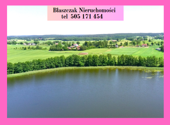 Zdjęcie z lotu ptaka przedstawia fragment jeziora z ciemną, spokojną taflą wody na pierwszym planie. Brzeg akwenu porasta równa, gęsta linia zielonych drzew liściastych, za którymi rozciągają się faliste, intensywnie zielone pola uprawne i łąki. W oddali, pośród drzew, widoczne są rozproszone zabudowania wiejskie oraz linia horyzontu zamknięta ścianą lasu. Całość kadru otoczona jest jasnoróżową ramką, a w górnej części umieszczono biały baner z czarnym napisem „Błaszczak Nieruchomości tel 505 171 454”.