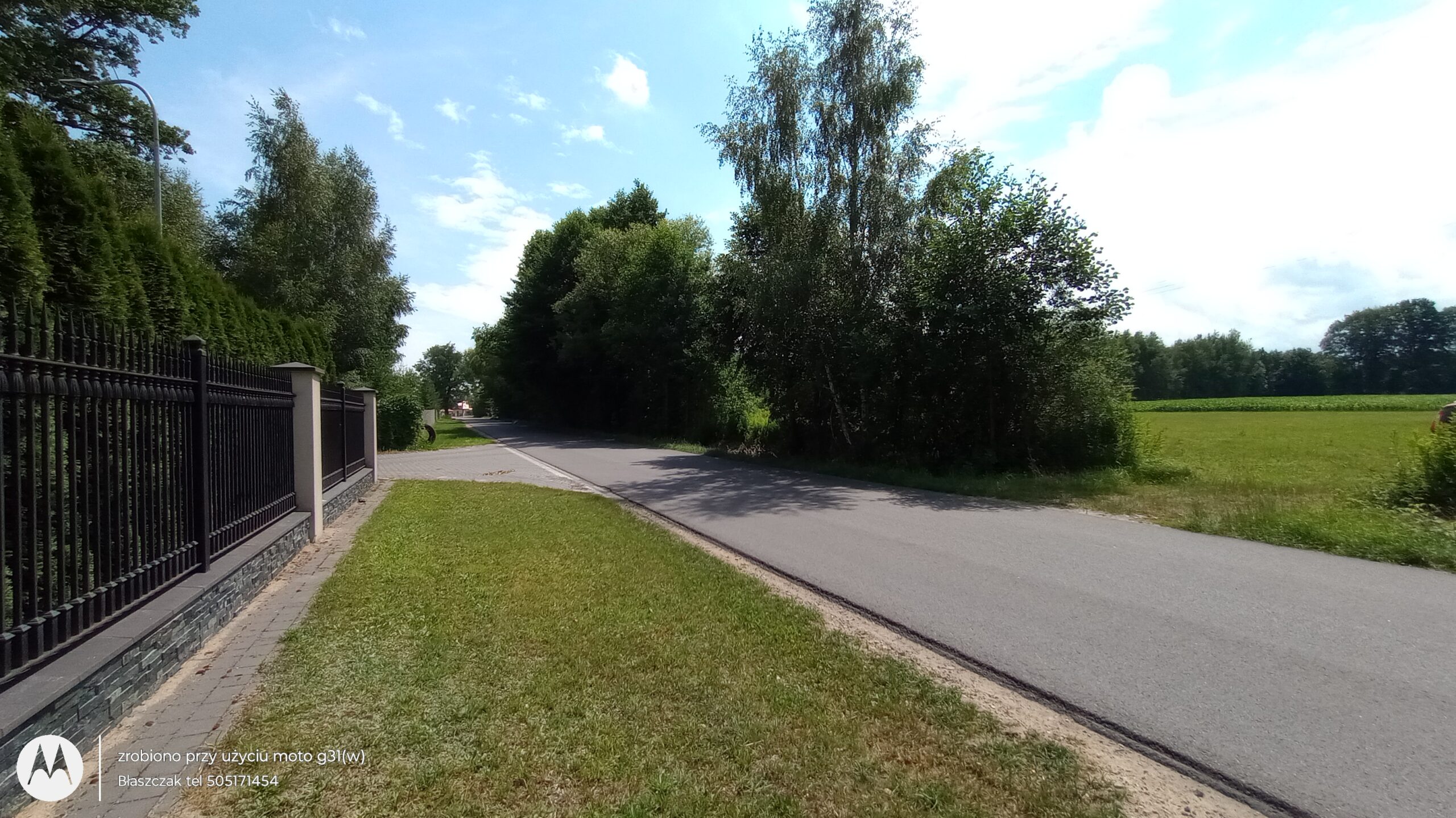Zdjęcie z poziomu gruntu przedstawia odcinek lokalnej, dwupasmowej drogi asfaltowej, która biegnie prosto w głąb kadru pod lekkim kątem. Po lewej stronie jezdni rozciąga się zadbany, szeroki pas zielonego trawnika, który graniczy z eleganckim ogrodzeniem posesji. Płot wykonany jest z czarnych, metalowych przęseł osadzonych na murowanej podmurówce z ozdobnego, ciemnego kamienia oraz jasnych słupkach, a tuż za nim pnie się gęsty, wysoki żywopłot z tui. Po prawej stronie drogi, na poboczu, rosną wysokie, bujne drzewa liściaste, które rzucają wyraźny cień na nawierzchnię szosy. Za linią drzew otwiera się widok na rozległe, zielone, uprawne pole. Nad całym krajobrazem roztacza się jasne, błękitne niebo z rozproszonymi, białymi chmurami, co wskazuje na słoneczny, letni dzień. W lewym dolnym rogu zdjęcia nałożono okrągłe, białe logo Motorola oraz napisy z danymi kontaktowymi agencji nieruchomości.