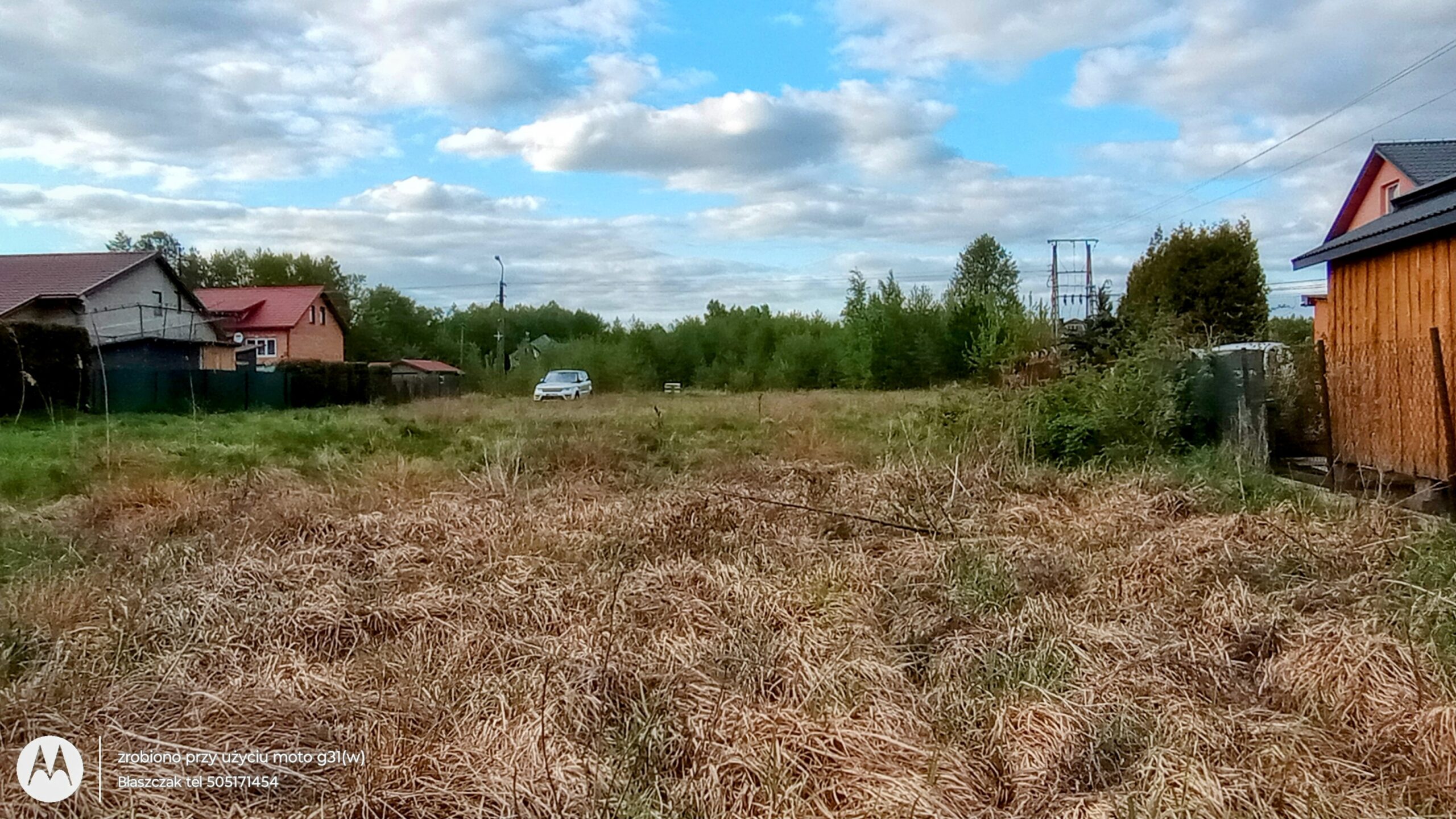 Zdjęcie z poziomu gruntu przedstawia otwartą, płaską działkę budowlaną lub pole. Na pierwszym planie dominuje gęsta, sucha, pożółkła trawa i zeszłoroczna roślinność łąkowa. W głębi terenu, pośrodku kadru, widać zaparkowany biały samochód osobowy. Po lewej stronie działki wznoszą się domy jednorodzinne o spadzistych, ciemnych i czerwonych dachach, odrodzone płotem. Po prawej stronie widoczny jest fragment drewnianego budynku gospodarczego lub ogrodzenia. Horyzont zamyka linia zielonych drzew i krzewów, nad którymi góruje konstrukcja słupa energetycznego. Nad całym krajobrazem rozciąga się błękitne niebo z rozproszonymi, białymi chmurami. W lewym dolnym rogu zdjęcia umieszczono subtelny, biały znak wodny.