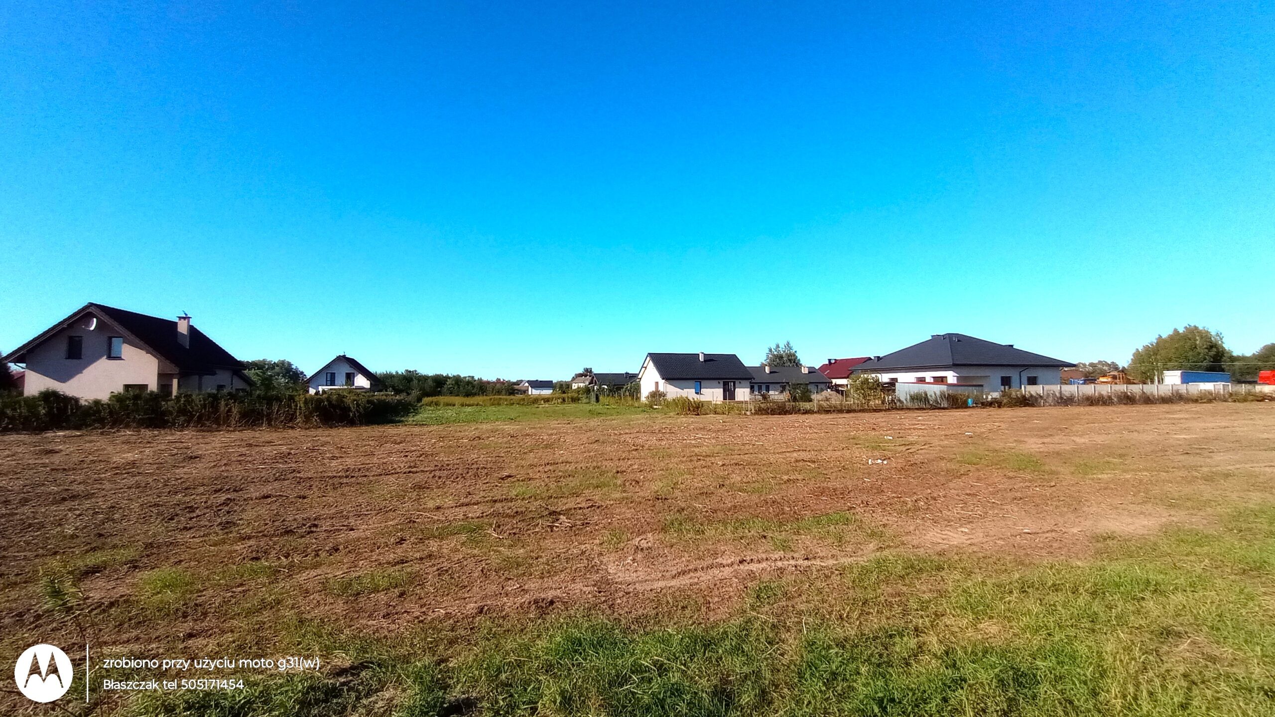 Fotografia przedstawia dużą, niezabudowaną działkę z widoczną suchą glebą i kępami zielonej trawy na pierwszym planie. Na horyzoncie, pod bezchmurnym, intensywnie błękitnym niebem, wznosi się rząd nowo wybudowanych domów jednorodzinnych o jasnych elewacjach i ciemnych, spadzistych dachach. W lewym dolnym rogu znajduje się jasny znak wodny aparatu oraz drobny tekst informacyjny z numerem telefonu.