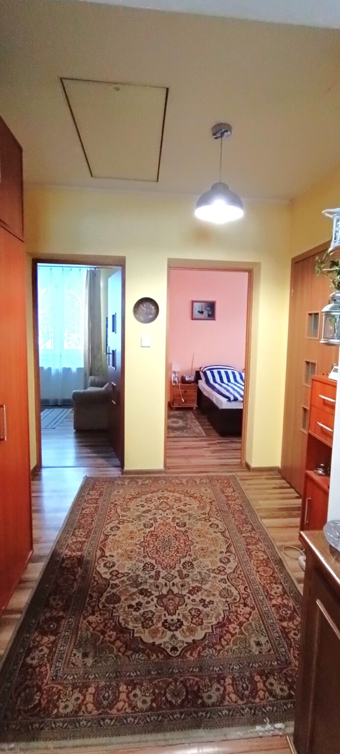Pionowe zdjęcie przedstawia długi korytarz lub przedpokój w mieszkaniu. Podłoga wyłożona jest panelami, na których leży duży, wzorzysty dywan w tradycyjne, orientalne motywy w odcieniach czerwieni, beżu i brązu. Ściany pomalowane są na jasny, żółty kolor, a u sufitu wisi nowoczesna lampa z metalowym, półkolistym kloszem. Na suficie widoczny jest również prostokątny właz na poddasze. W głębi korytarza znajdują się dwa otwarte przejścia do innych pomieszczeń: po lewej stronie widać fragment salonu z kanapą i oknem z firanką, a po prawej sypialnię z łóżkiem zasłanym pasiastą pościelą i szafką nocną. Po bokach korytarza stoją drewniane meble oraz widoczne są drzwi wewnętrzne.