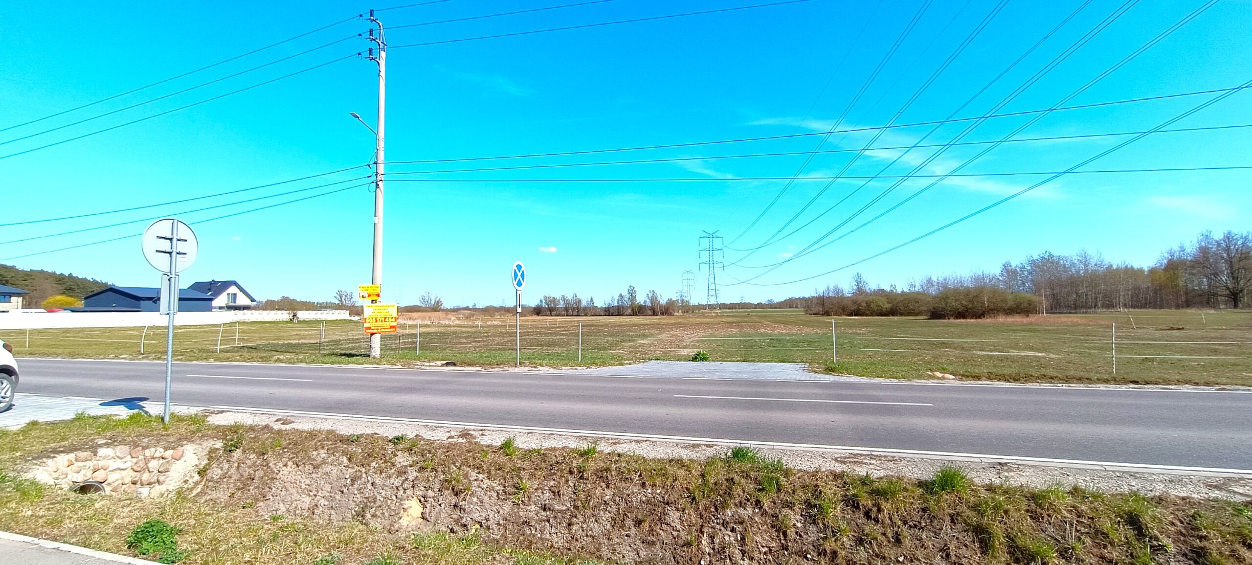Zdjęcie z poziomu gruntu przedstawia szerokie ujęcie lokalnej drogi asfaltowej biegnącej poziomo przez środek kadru. Na pierwszym planie widoczne jest trawiaste pobocze z ziemnym rowem melioracyjnym, w którym po lewej stronie znajduje się kamienne umocnienie przepustu. Nad rowem stoi pionowy znak drogowy. Po drugiej stronie jezdni rozciąga się płaski, rozległy teren zielony z rzadką roślinnością, zabezpieczony prowizorycznym ogrodzeniem z drutu. Na działkę prowadzi utwardzony, szary zjazd. Po lewej stronie stoi wysoki betonowy słup energetyczny z żółtą tablicą reklamową, od którego odchodzą przewody. W tle, po prawej stronie, nad horyzontem dominują potężne, stalowe słupy kratownicowe napowietrznej linii wysokiego napięcia. Nad całością rozciąga się bezchmurne, intensywnie błękitne niebo w pełnym słońcu.