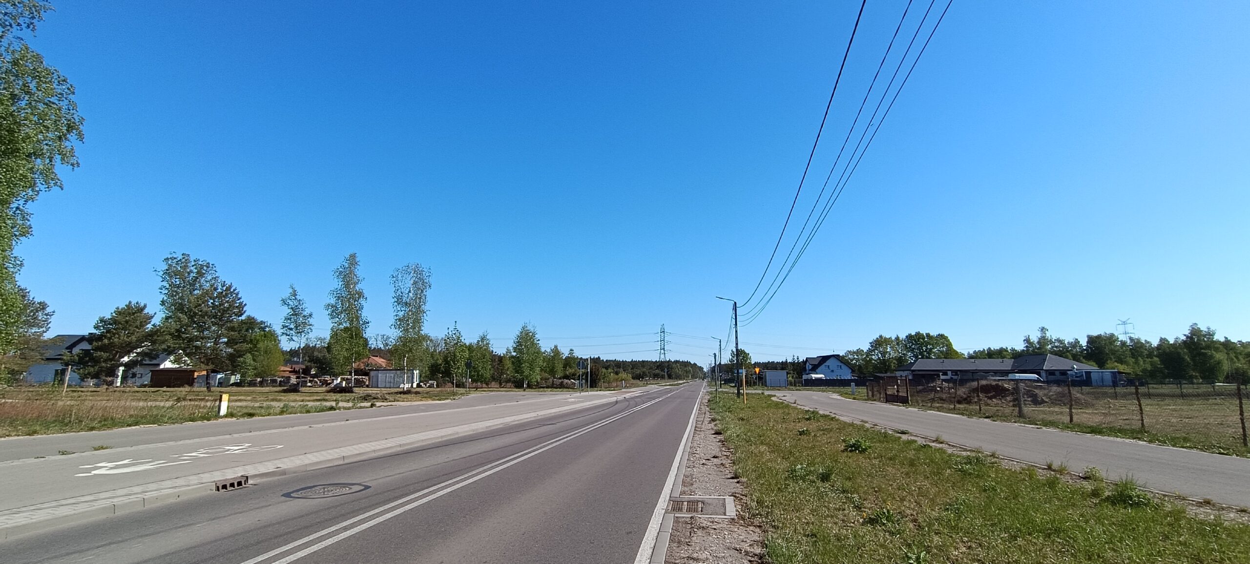 Widok na asfaltową drogę przebiegającą w pobliżu nieruchomości, z zabudową jednorodzinną, terenami zielonymi oraz infrastrukturą drogową w spokojnej okolicy.