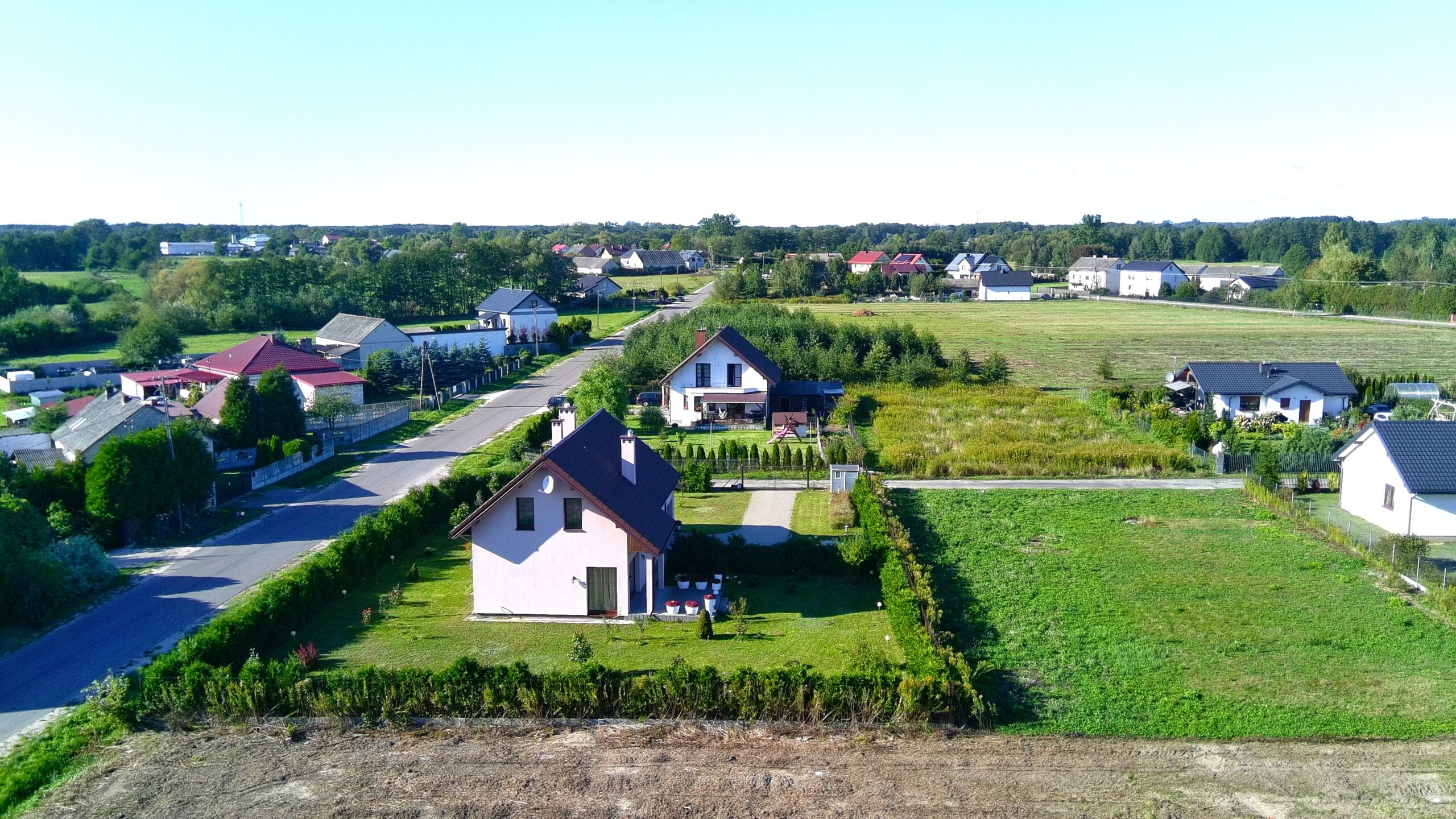Fotografia wykonana z lotu ptaka przedstawia kameralne osiedle domów jednorodzinnych w otoczeniu zieleni. Na pierwszym planie, w dolnej części kadru, widoczny jest dom z jasną elewacją i ciemnym, spadzistym dachem, otoczony zadbanym ogrodem i gęstym żywopłotem. Po lewej stronie w głąb obrazu biegnie lokalna droga asfaltowa, przy której usytuowane są kolejne posesje. Po prawej stronie oraz w tle rozciągają się zielone, niezabudowane działki oraz otwarte pola przechodzące w linię lasu pod czystym, błękitnym niebem.