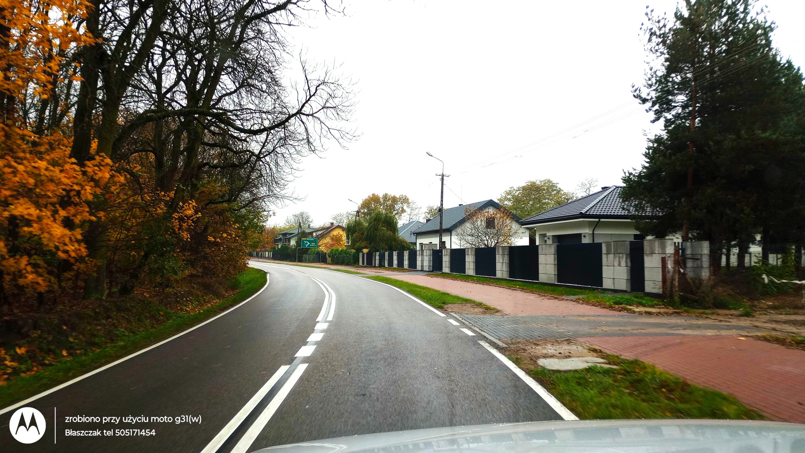 Zdjęcie zrobione z wnętrza pojazdu (w dolnej części widoczny jest fragment maski samochodu) przedstawia łagodny zakręt dwupasmowej, wilgotnej drogi asfaltowej z wyraźnym, białym oznakowaniem poziomym. Po lewej stronie szosy ciągnie się gęsta ściana lasu z drzewami o bezlistnych koronach oraz krzewami o intensywnie żółtych i pomarańczowych, jesiennych liściach. Po prawej stronie drogi przebiega chodnik z czerwonej kostki brukowej, za którym wznosi się nowoczesne, grafitowo-szare ogrodzenie z betonowych bloczków i paneli. Za płotem stoją nowe, parterowe domy jednorodzinne z ciemnymi, spadzistymi dachami. Nad całością roztacza się jednolicie jasne, zachmurzone niebo, a w lewym dolnym rogu nałożone jest okrągłe logo marki Motorola oraz biały tekst informacyjny agencji nieruchomości.